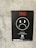A black poster is attached to a concrete wall. The poster features a sad face symbol with a disposable cup and bottle inside, emphasizing the negative impact of single-use items. The text 'Single use, Think twice' is prominently displayed in red above the face. Additional information and logos, including a QR code, are at the bottom.
