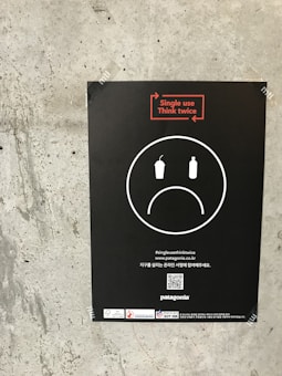 A black poster is attached to a concrete wall. The poster features a sad face symbol with a disposable cup and bottle inside, emphasizing the negative impact of single-use items. The text 'Single use, Think twice' is prominently displayed in red above the face. Additional information and logos, including a QR code, are at the bottom.