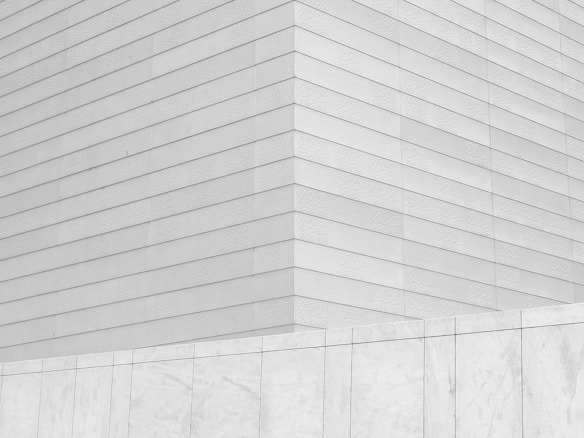 A minimalist architectural view featuring a geometric pattern of horizontal lines on a building's facade. The structure is composed of uniform, smooth panels creating a sleek, modern appearance. A portion of the image includes a low wall in a slightly lighter shade, adding contrast.
