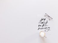 A minimalistic setup featuring a white background with a piece of paper containing handwritten black text that reads 'joy comes in the morning.' A pair of glasses rests beside the paper, and there is a cup of frothy coffee positioned at the bottom.