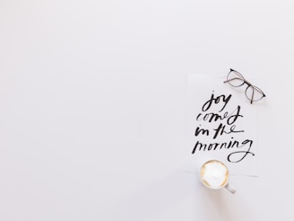 A minimalistic setup featuring a white background with a piece of paper containing handwritten black text that reads 'joy comes in the morning.' A pair of glasses rests beside the paper, and there is a cup of frothy coffee positioned at the bottom.