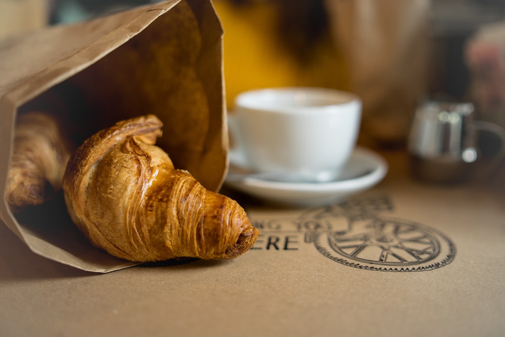 baked bread on paper bag beside white cup and saucer