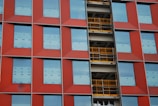 The image displays a building facade featuring a grid pattern with red panels and large rectangular windows. In the middle, there is a section with exposed yellow construction railings and unfinished surfaces.