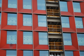 The image displays a building facade featuring a grid pattern with red panels and large rectangular windows. In the middle, there is a section with exposed yellow construction railings and unfinished surfaces.