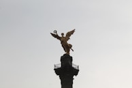 A golden angelic statue stands atop a tall, ornate column against a pale sky. The statue is in a dynamic pose, holding a laurel wreath in one hand.