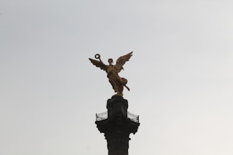 A golden angelic statue stands atop a tall, ornate column against a pale sky. The statue is in a dynamic pose, holding a laurel wreath in one hand.