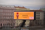 A large outdoor billboard displaying an advertisement with a nutcracker image and text in Czech. The billboard is situated above a busy city street, and a row of classical-style buildings is visible in the background, including a pink building labeled as a hotel.