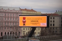 A large outdoor billboard displaying an advertisement with a nutcracker image and text in Czech. The billboard is situated above a busy city street, and a row of classical-style buildings is visible in the background, including a pink building labeled as a hotel.