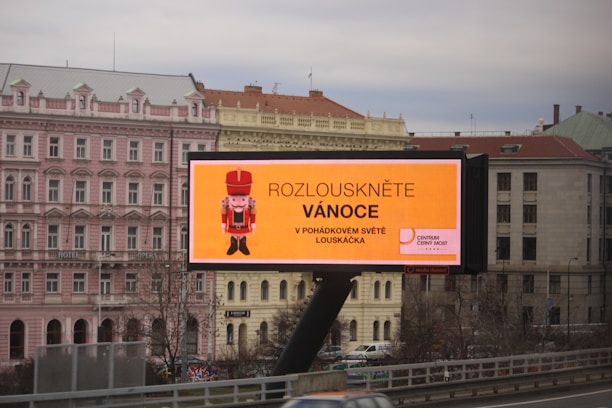 A large outdoor billboard displaying an advertisement with a nutcracker image and text in Czech. The billboard is situated above a busy city street, and a row of classical-style buildings is visible in the background, including a pink building labeled as a hotel.
