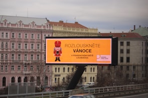 A large outdoor billboard displaying an advertisement with a nutcracker image and text in Czech. The billboard is situated above a busy city street, and a row of classical-style buildings is visible in the background, including a pink building labeled as a hotel.