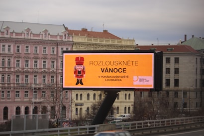 A large outdoor billboard displaying an advertisement with a nutcracker image and text in Czech. The billboard is situated above a busy city street, and a row of classical-style buildings is visible in the background, including a pink building labeled as a hotel.