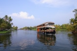 A traditional houseboat navigating through a serene waterway, surrounded by lush greenery and palm trees. The blue sky has scattered clouds and the water reflects the natural scenery.