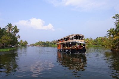 A traditional houseboat navigating through a serene waterway, surrounded by lush greenery and palm trees. The blue sky has scattered clouds and the water reflects the natural scenery.