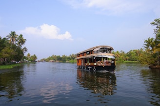 A traditional houseboat navigating through a serene waterway, surrounded by lush greenery and palm trees. The blue sky has scattered clouds and the water reflects the natural scenery.