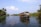 A traditional houseboat navigating through a serene waterway, surrounded by lush greenery and palm trees. The blue sky has scattered clouds and the water reflects the natural scenery.