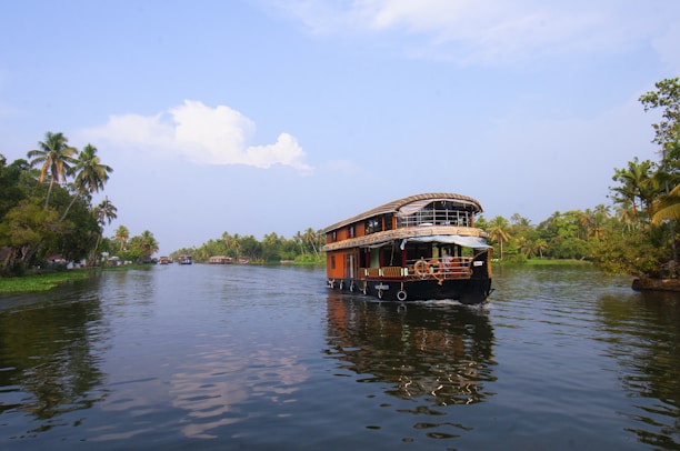 A traditional houseboat navigating through a serene waterway, surrounded by lush greenery and palm trees. The blue sky has scattered clouds and the water reflects the natural scenery.