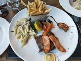A white plate features a diverse meal with pasta in a creamy sauce, a piece of grilled steak topped with salmon slices, and a selection of vegetables including broccoli, carrot sticks, and a slice of corn. A small serving of baked beans is placed in a spoon, and a slice of lemon is on the side. A metal basket holds a portion of French fries.