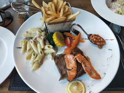 A white plate features a diverse meal with pasta in a creamy sauce, a piece of grilled steak topped with salmon slices, and a selection of vegetables including broccoli, carrot sticks, and a slice of corn. A small serving of baked beans is placed in a spoon, and a slice of lemon is on the side. A metal basket holds a portion of French fries.