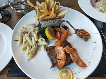 A white plate features a diverse meal with pasta in a creamy sauce, a piece of grilled steak topped with salmon slices, and a selection of vegetables including broccoli, carrot sticks, and a slice of corn. A small serving of baked beans is placed in a spoon, and a slice of lemon is on the side. A metal basket holds a portion of French fries.