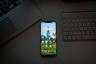 A smartphone placed on a desk between a laptop and an external keyboard. The phone's screen is on and displays various app icons against a background image of a building and trees.