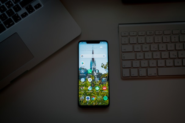 A smartphone placed on a desk between a laptop and an external keyboard. The phone's screen is on and displays various app icons against a background image of a building and trees.