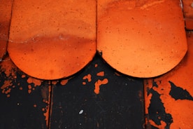 A close-up view of two overlapping, weathered orange ceramic tiles with visible dirt and speckled texture. These tiles lie on a black surface with orange paint chipping away, revealing the contrasting color underneath.