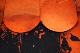 A close-up view of two overlapping, weathered orange ceramic tiles with visible dirt and speckled texture. These tiles lie on a black surface with orange paint chipping away, revealing the contrasting color underneath.