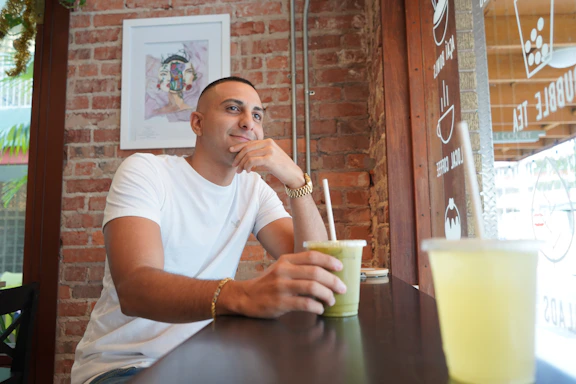 A softly lit photo of a stylish woman holding a vibrant green juice in a cozy, neutral-toned cafe setting with leafy plants and light wood accents.