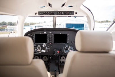 The image depicts the interior cockpit of a small aircraft, featuring two pilot seats and a control panel with various instruments and screens. The view through the front window shows a blurred outdoor area with a building in the background.