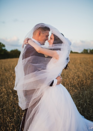 A couple is embracing closely in a wheat field, enveloped in flowing white fabric. The groom is in a dark suit and the bride is in a flowing white wedding dress. The scene captures a serene and romantic moment during sunset, with a soft blue sky and scattered clouds in the background.