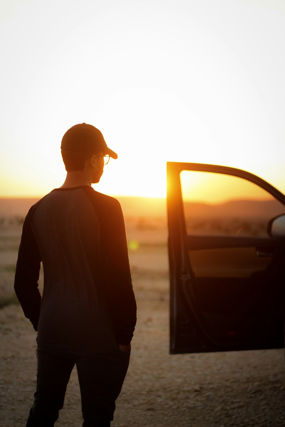 man standing beside car with open door facing setting sun