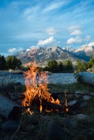 A campfire with vibrant flames burns on a grassy area surrounded by stones, with a backdrop of majestic snow-capped mountains and a blue sky dotted with clouds.