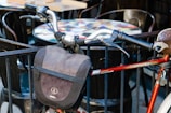 A close-up of a bicycle handlebar adorned with a black bag, set against a background of outdoor café tables and chairs. The bicycle frame is red, and the handle grips are black, showing signs of wear.