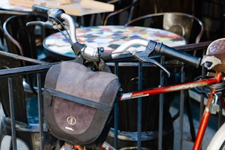 A close-up of a bicycle handlebar adorned with a black bag, set against a background of outdoor café tables and chairs. The bicycle frame is red, and the handle grips are black, showing signs of wear.