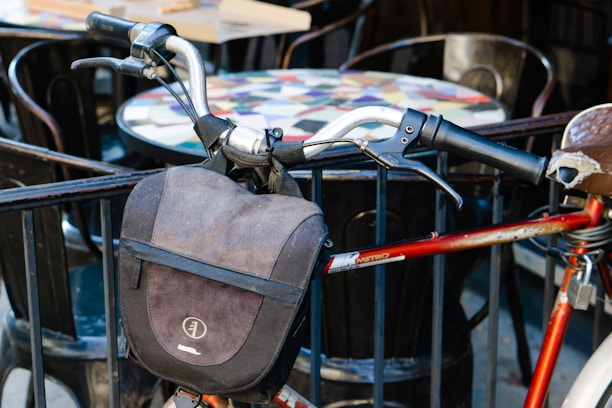 A close-up of a bicycle handlebar adorned with a black bag, set against a background of outdoor café tables and chairs. The bicycle frame is red, and the handle grips are black, showing signs of wear.