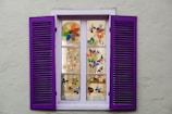 A window with purple shutters, featuring multiple panes displaying colorful stained glass designs of flowers and butterflies. The window is set in a light gray wall, with each pane showcasing intricate artwork that enhances the aesthetic appeal.