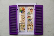 A window with purple shutters, featuring multiple panes displaying colorful stained glass designs of flowers and butterflies. The window is set in a light gray wall, with each pane showcasing intricate artwork that enhances the aesthetic appeal.