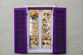 A window with purple shutters, featuring multiple panes displaying colorful stained glass designs of flowers and butterflies. The window is set in a light gray wall, with each pane showcasing intricate artwork that enhances the aesthetic appeal.