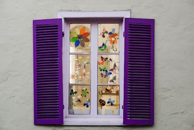 A window with purple shutters, featuring multiple panes displaying colorful stained glass designs of flowers and butterflies. The window is set in a light gray wall, with each pane showcasing intricate artwork that enhances the aesthetic appeal.