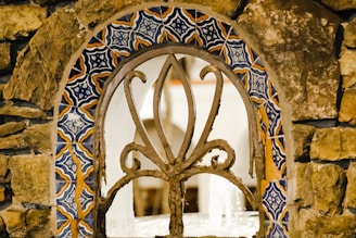 A decorative stone wall featuring a wrought iron grate with intricate patterns. Surrounding the grate, there are colorful ceramic tiles in blue, orange, and white, creating a striking visual contrast with the natural stone.