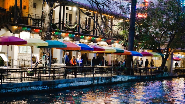 A riverside restaurant features tables with colorful umbrellas in an outdoor seating area. Twinkling string lights add a festive atmosphere, while reflections create a vibrant pattern on the water below. People are casually gathered, enhancing the lively ambiance.