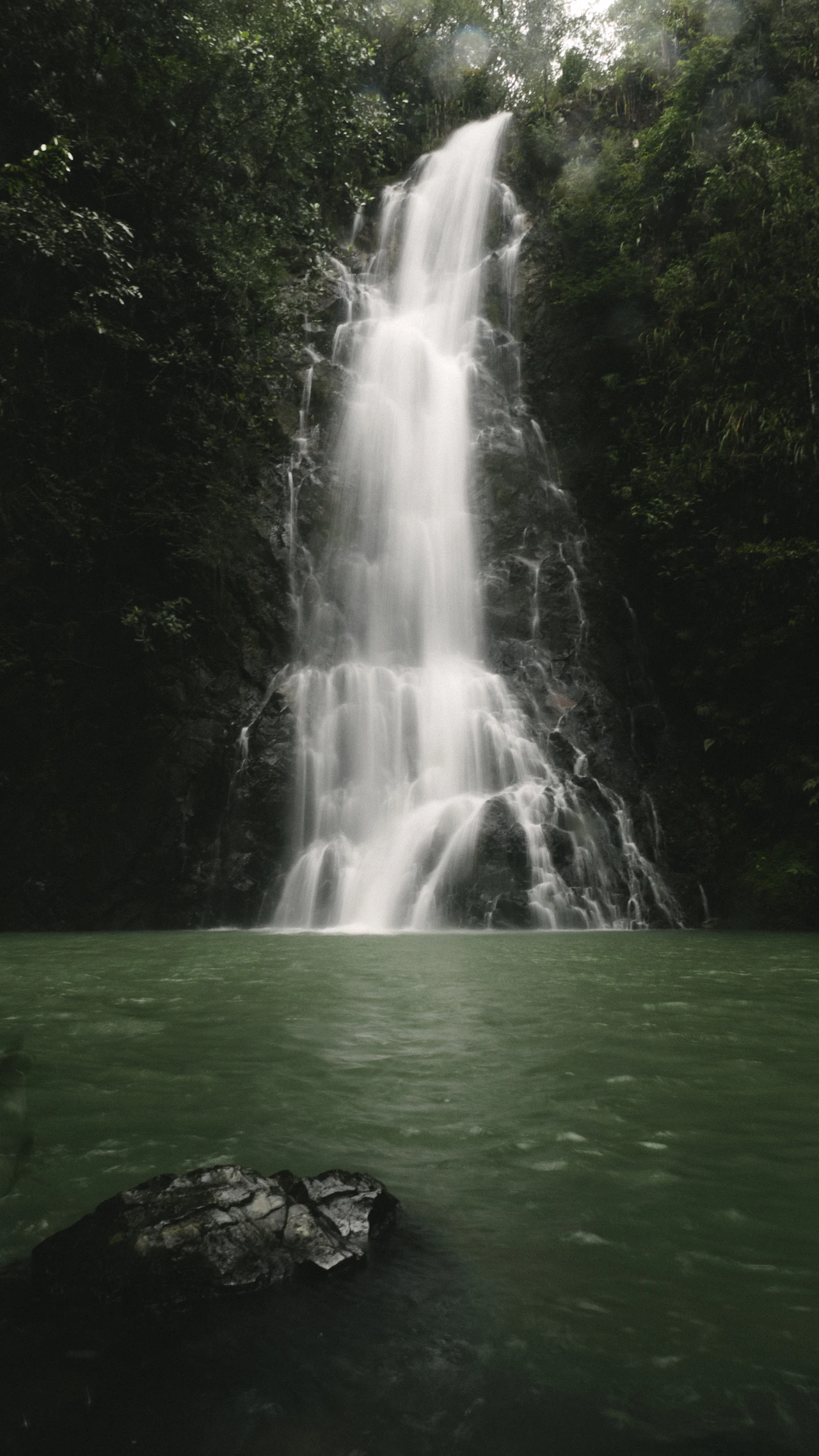A tall, narrow waterfall dropping down a dark rock face into a deep green pool, framed by forest.