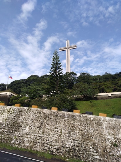 A large cross monument stands prominently against a backdrop of a cloudy blue sky. Below the cross, lush greenery covers a hillside, and a textured stone wall with yellow and black markers runs across the foreground. A flagpole with a flag fluttering is visible to the left.
