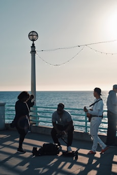 People are gathered on a pier by the ocean during a sunny day. A musician plays a guitar while others listen nearby. The scene is illuminated by warm sunlight, casting long shadows on the ground. A string of lights hangs from a lamp post, adding a decorative touch.