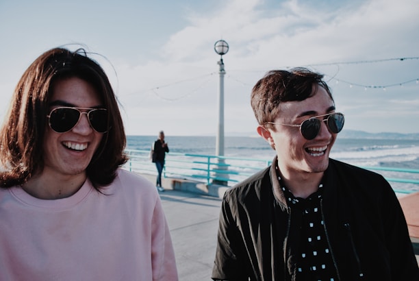 Two people laughing together while seated side-by-side on a sleek paddleboard catamaran near a sandy beach.