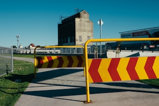 A metal barrier with red and yellow chevron stripes blocks a paved pathway. In the background, an industrial building with a slanted roof and a billboard are visible. A chain-link fence and grassy areas are on either side of the path.