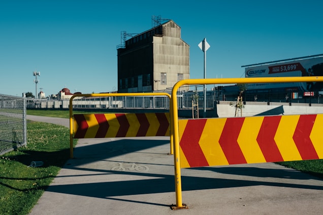 A metal barrier with red and yellow chevron stripes blocks a paved pathway. In the background, an industrial building with a slanted roof and a billboard are visible. A chain-link fence and grassy areas are on either side of the path.