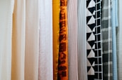 A variety of fabric panels hanging side by side, showcasing different patterns and colors. The fabrics include solid colors, stripes, geometric patterns, and abstract designs.