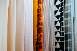 A variety of fabric panels hanging side by side, showcasing different patterns and colors. The fabrics include solid colors, stripes, geometric patterns, and abstract designs.