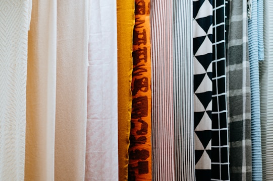 A variety of fabric panels hanging side by side, showcasing different patterns and colors. The fabrics include solid colors, stripes, geometric patterns, and abstract designs.